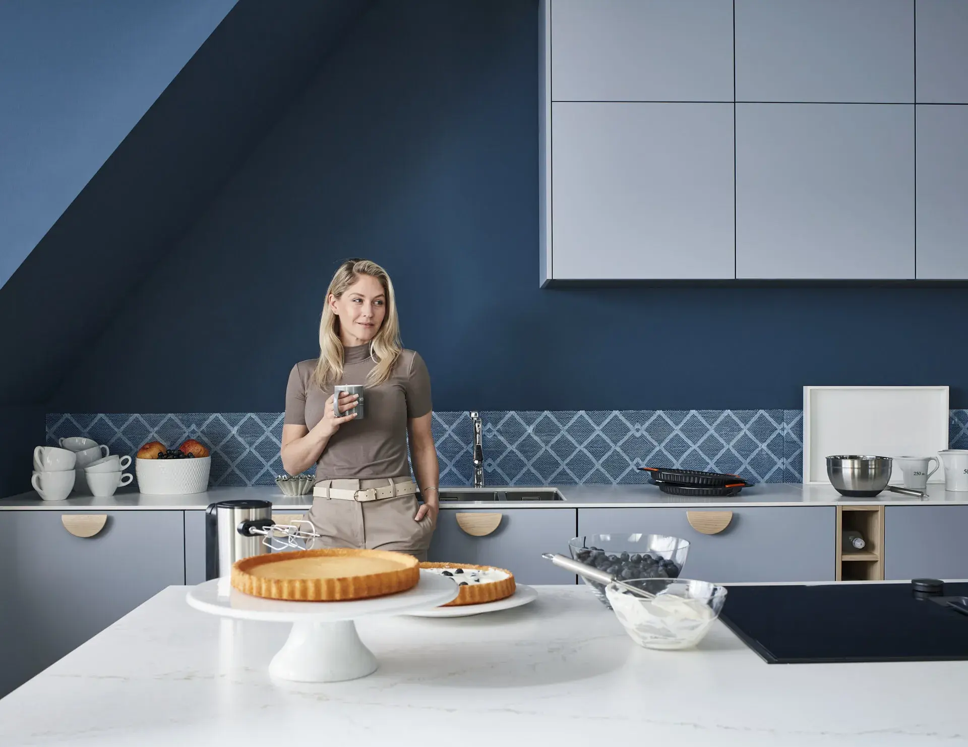 Model lehnt sich an die Edelstahlspüle, Kücheninsel mit NEFF Induktionskochfeld mit Muldenlüfter, Schublade, Mix aus Lack, Matt und Eiche